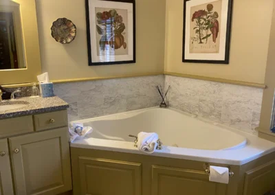 A bathroom with a corner bathtub, beige cabinetry, and botanical framed art.