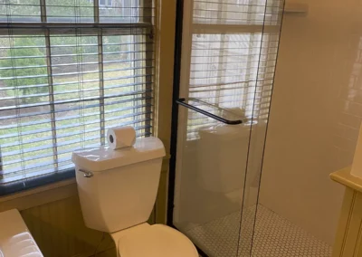 A bathroom with a toilet, glass shower stall, and a window with blinds.